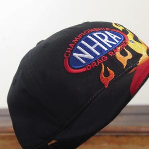 May include: Black baseball cap with embroidered "NHRA Drag Racing Championship" logo in white, red, and blue. Flames in red, yellow, and orange adorn the side and brim of the hat.