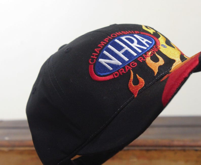 May include: Black baseball cap with embroidered "NHRA Drag Racing Championship" logo in white, red, and blue. Flames in red, yellow, and orange adorn the side and brim of the hat.