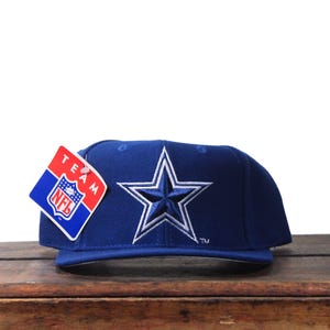 May include: A navy blue baseball cap with a white star emblem. The cap has a flat peak and a tag with the NFL logo. The cap is displayed on a wooden surface.