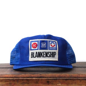 May include: A blue trucker hat with a mesh back and a rectangular patch. The patch features the text "BLANKENSHIP" and logos for AC, GM, and Delco. The hat is sitting on a wooden surface.