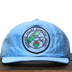 Puede incluir: Una gorra de camionero de malla azul claro con un parche blanco y negro que dice "Mohave Valley Athletic Assoc." con un gráfico de un campo de béisbol, una cancha de baloncesto y un campo de fútbol.