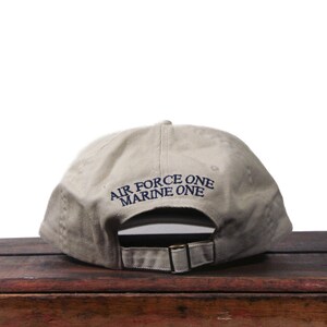 Puede incluir: Gorra de b&eacute;isbol caqui con el texto azul marino "AIR FORCE ONE MARINE ONE". La gorra tiene una correa ajustable y una visera curva. El sombrero est&aacute; sobre una superficie de madera.