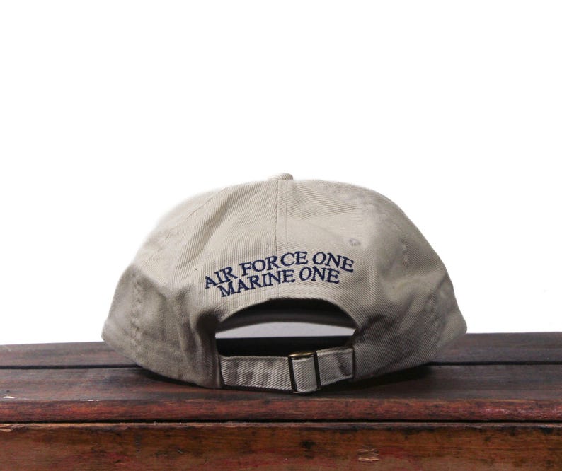 Puede incluir: Gorra de b&eacute;isbol caqui con el texto azul marino "AIR FORCE ONE MARINE ONE". La gorra tiene una correa ajustable y una visera curva. El sombrero est&aacute; sobre una superficie de madera.