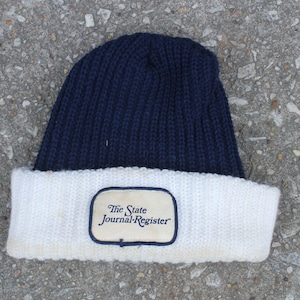 以下が含まれることがあります： 紺色と白のニットのビーニー。白いパッチには「The State Journal-Register」と書いてあります。