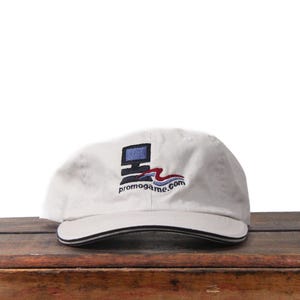 Puede incluir: Gorra de béisbol color crema con borde a rayas blancas y negras. La gorra presenta un logotipo bordado de un monitor de computadora y el texto "promogame.com" en azul, rojo y negro.