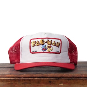 May include: A white and red Pac-Man trucker hat. The hat has a white front panel with a Pac-Man logo and a red brim and mesh back. The Pac-Man logo includes the text "PAC-MAN" in yellow and the Midway logo.