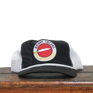 Puede incluir: Una gorra de camionero negra y blanca con un parche rojo, blanco y amarillo que dice "Army TACMS The Decisive Edge".