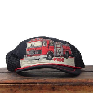 Può includere: Cappellino da camionista nero con un disegno di un'autopompa rossa. L'autopompa ha dettagli argentati e il testo "FMC" è visibile. Il cappello ha un retro in rete nera e un bordo rosso.