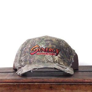 May include: Camouflage baseball cap with the text "SYNERGY TECHNOLOGIES" in red and orange. The cap has a distressed brim and a brown back panel. The hat is sitting on a wooden surface.