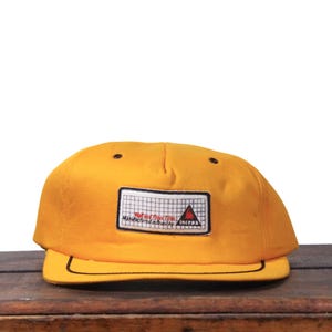 May include: Mustard yellow baseball cap with a black brim and a rectangular patch. The patch features a grid pattern, text, and a red triangle. The cap is sitting on a wooden surface.