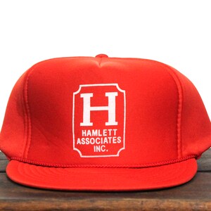 Könnte beinhalten: Rote Trucker-Kappe mit einem weißen Logo, das "HAMLETT ASSOCIATES INC." in einem rechteckigen Rahmen mit einem großen weißen "H" in der Mitte zeigt.