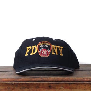 Op de afbeelding: Zwarte baseballcap met gouden "FDNY" letters. De cap heeft een patch met de tekst "Fire Department City of New York". De rand heeft een witte en grijze rand.
