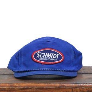 Pode incluir: Um boné azul com o texto "SCHMIDT BAKING COMPANY" num patch oval vermelho e branco. O boné tem um detalhe de corda ao longo da aba e está sobre uma superfície de madeira.