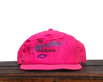 May include: A pink baseball cap with a blue and white embroidered logo that says "The Heartbeat of America".