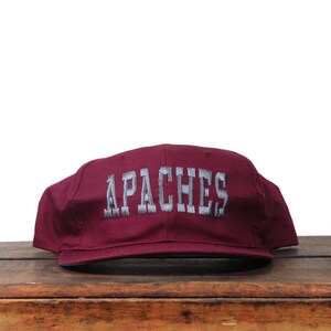 Op de afbeelding: Bordeauxrode baseballpet met het woord "APACHES" in grijze blokletters geborduurd. De pet ligt op een houten oppervlak tegen een witte achtergrond.