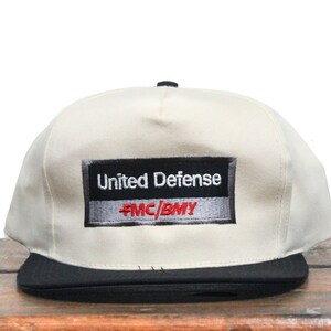 Può includere: Un cappello da baseball bianco e nero con visiera nera. La parte anteriore del cappello ha una toppa nera e grigia con il testo "United Defense" in bianco e "FMC/BMY" in rosso.