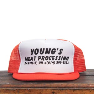 Könnte beinhalten: Eine weiße und orangefarbene Trucker-Kappe mit dem Aufdruck "YOUNG'S MEAT PROCESSING DANVILLE, OH (614) 599-6833" auf der Vorderseite.