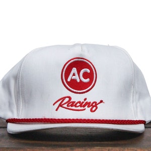 May include: White baseball cap with a red circle logo that says "AC" and the text "Racing" in red cursive.