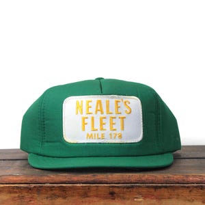 Puede incluir: Gorra de camionero verde con visera plana. La gorra presenta un parche rectangular blanco con las palabras "NEALE'S FLEET" en letras doradas y "MILE 178" debajo. La gorra está sobre una superficie de madera.