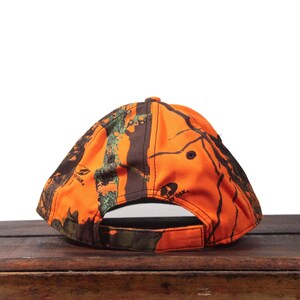 May include: A camouflage baseball cap in shades of orange, brown, and green. The cap features a curved brim and an adjustable strap at the back. The cap has the Mossy Oak logo on the side.