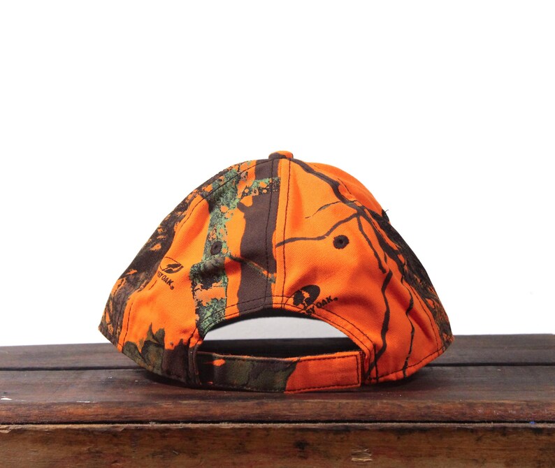 May include: A camouflage baseball cap in shades of orange, brown, and green. The cap features a curved brim and an adjustable strap at the back. The cap has the Mossy Oak logo on the side.