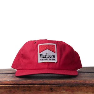 May include: A red baseball cap with a white and red Marlboro Racing Team logo patch. The cap is made of a sturdy material and has a curved brim. The hat is sitting on a wooden surface.