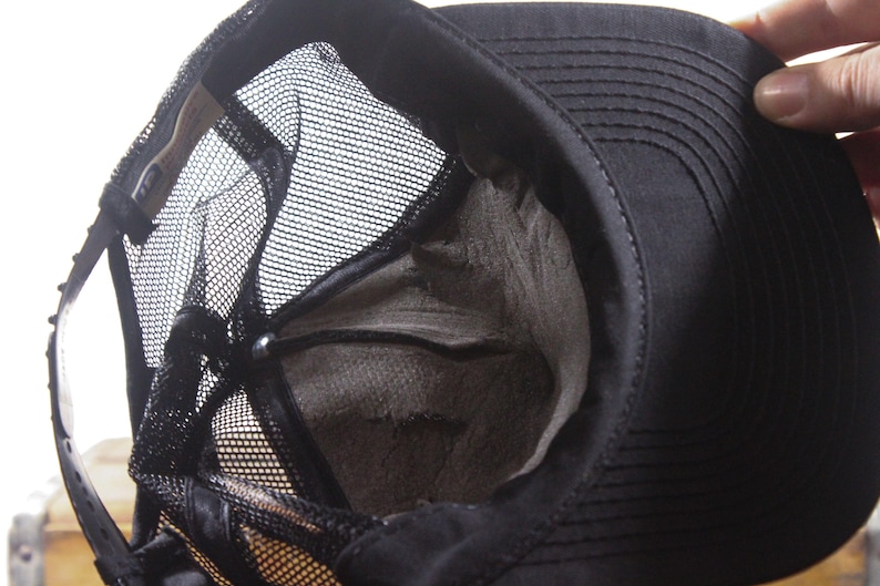 May include: Black trucker hat with a mesh back and a structured front. The hat's interior is visible, showing a worn, gray lining. The brim is curved, and the hat appears to be made of durable fabric.
