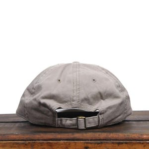 Puede incluir: Una gorra de b&eacute;isbol de algod&oacute;n gris con una correa ajustable marr&oacute;n. La gorra est&aacute; sobre una superficie de madera.