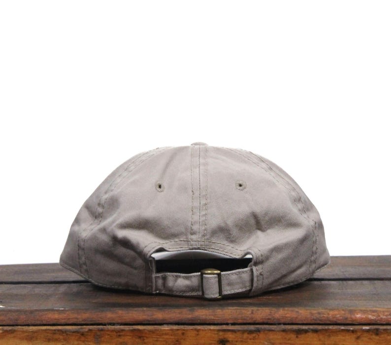 Puede incluir: Una gorra de b&eacute;isbol de algod&oacute;n gris con una correa ajustable marr&oacute;n. La gorra est&aacute; sobre una superficie de madera.
