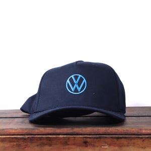 May include: A navy blue baseball cap featuring a light blue embroidered Volkswagen logo on the front. The cap has a curved brim and a structured crown. The hat is displayed on a wooden surface.