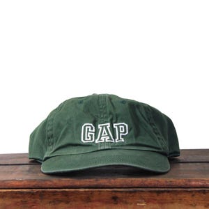 May include: A dark green baseball cap with the word "GAP" embroidered in white block letters. The cap is made of a soft material and has a curved brim. The hat is sitting on a wooden surface.