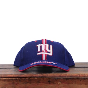 Könnte beinhalten: Eine marineblaue Baseballkappe mit den weiß und rot gestickten Buchstaben ny auf der Vorderseite und einem vertikalen grauen und roten Streifen in der Mitte. Der Schirm ist mit einem roten Rand und dem in Rot gestickten Wort Giants versehen.