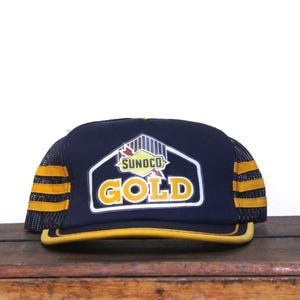 Puede incluir: Gorra de camionero azul marino y amarilla con el texto "SUNOCO GOLD" en un diseño blanco y amarillo. La gorra tiene una parte trasera de malla y rayas amarillas en los laterales.