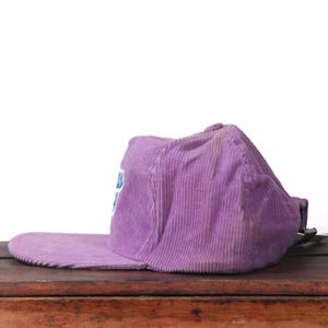 May include: A purple corduroy baseball cap with a flat brim. The cap has a faded appearance and a small logo on the front. The hat is resting on a wooden surface.