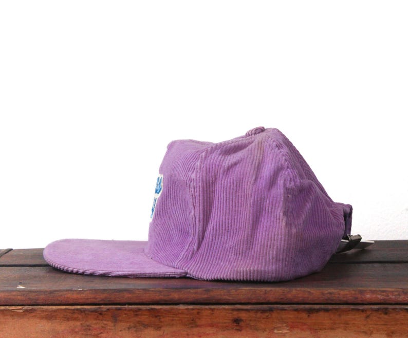 May include: A purple corduroy baseball cap with a flat brim. The cap has a faded appearance and a small logo on the front. The hat is resting on a wooden surface.