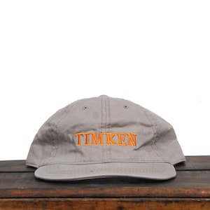 Puede incluir: Una gorra de b&eacute;isbol gris con un logotipo bordado en naranja que dice "TIMKEN".