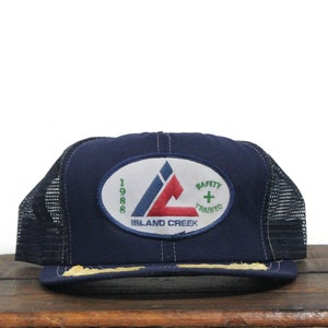 Puede incluir: Una gorra de camionero de malla azul y negra con un parche blanco que presenta un logotipo triangular rojo y azul con el texto "1988 Island Creek Safety + Trained".