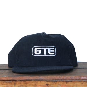 May include: Black corduroy baseball cap with a flat brim. The cap features the white embroidered letters "GTE" inside a white rectangular border. The hat is displayed on a wooden surface against a white background.