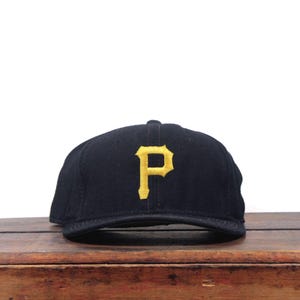 May include: A black baseball cap featuring a prominent, embroidered yellow letter P centred on the front panel. The cap has a structured crown and a curved brim, showcasing a classic design with clean stitching and a solid dark fabric construction.