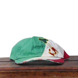 May include: A green, white, and red baseball cap with an embroidered design. The cap features the colors of the Mexican flag. The cap is made of a soft material and is sitting on a wooden surface.