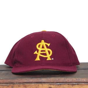 Op de afbeelding: Bordeaux baseballpet met een goud geborduurd "AS" logo. De pet heeft een gebogen klep en een gele onderkant. De hoed ligt op een houten oppervlak.