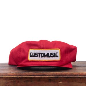 Puede incluir: Una gorra de béisbol de color rojo brillante con un parche rectangular en la parte delantera. El parche tiene un fondo blanco con un borde amarillo y el texto "CUSTOMUSIC" bordado en letras mayúsculas negras en el centro.