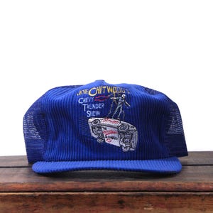 May include: A blue corduroy trucker hat with a mesh back. The hat features an embroidered design of a car and a stuntman, with the text "JOIE CHITWOOD'S CHEVY THUNDER SHOW".