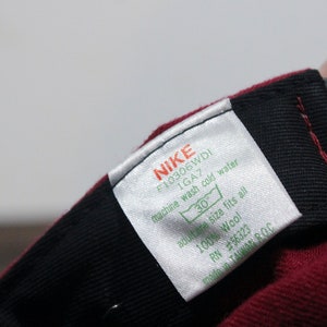 May include: A white tag with black text that reads "NIKE F10306WDI 1GA7 machine wash cold water 30 adult one size fits all 100% Wool RN #56323 made in TAIWAN ROC". The tag is attached to a maroon baseball cap.