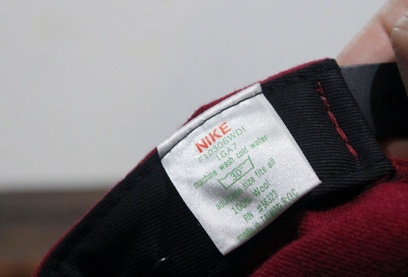 May include: A white tag with black text that reads "NIKE F10306WDI 1GA7 machine wash cold water 30 adult one size fits all 100% Wool RN #56323 made in TAIWAN ROC". The tag is attached to a maroon baseball cap.