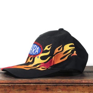 May include: Black baseball cap with embroidered flames in yellow, orange, and red. The cap features the NHRA Racing logo on the side. The brim is red.