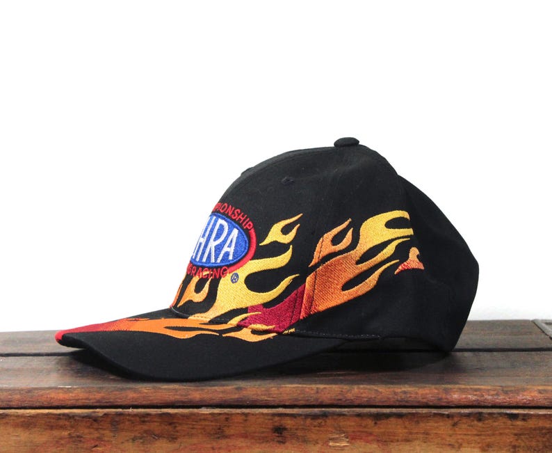 May include: Black baseball cap with embroidered flames in yellow, orange, and red. The cap features the NHRA Racing logo on the side. The brim is red.