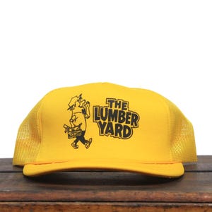 May include: A yellow trucker hat with a black mesh back. The hat has a black and white illustration of a person carrying wood and the text "The Lumber Yard" in black.