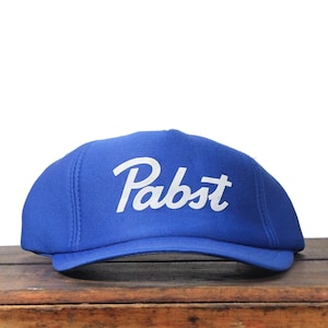 Puede incluir: Gorra de béisbol azul con la palabra "Pabst" en blanco en una fuente de escritura. La gorra está sobre una superficie de madera. El fondo es blanco.