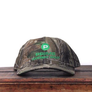 May include: Camouflage baseball cap with the text "PRIME EQUIPMENT GROUP" embroidered in green. The cap features a green "P" logo. The hat is made of a camouflage pattern fabric.